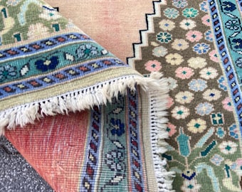 May include: Close-up of a section of a vintage rug with intricate patterns. The rug features a palette of colors including teal, pink, and brown. The design includes floral and geometric motifs, with a fringed edge.