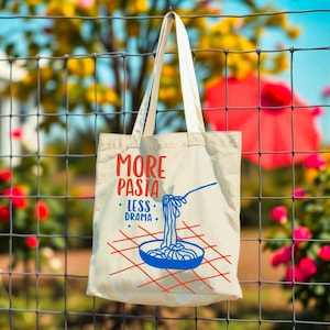 Puede incluir: Bolso tote de color crema con el texto "MORE PASTA LESS DRAMA" en rojo y azul. El dise&ntilde;o incluye un cuenco azul de pasta con una cuchara y un patr&oacute;n de cuadr&iacute;cula roja. La bolsa est&aacute; colgada de una valla de alambre.