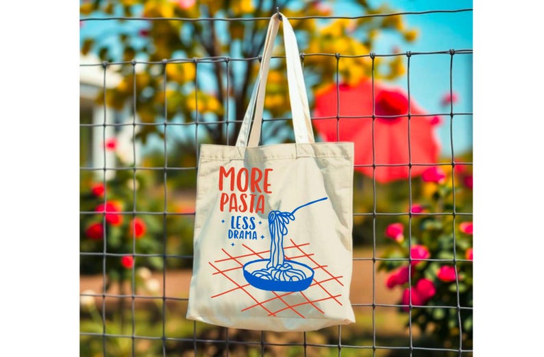 Puede incluir: Bolso tote de color crema con el texto "MORE PASTA LESS DRAMA" en rojo y azul. El dise&ntilde;o incluye un cuenco azul de pasta con una cuchara y un patr&oacute;n de cuadr&iacute;cula roja. La bolsa est&aacute; colgada de una valla de alambre.