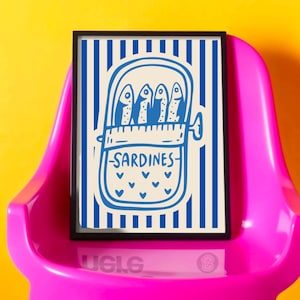 Puede incluir: Una impresi&oacute;n art&iacute;stica enmarcada con una ilustraci&oacute;n azul y blanca de sardinas enlatadas. La obra de arte presenta un fondo a rayas y la palabra "Sardines" escrita debajo. La impresi&oacute;n se muestra en una silla rosa sobre un fondo amarillo.