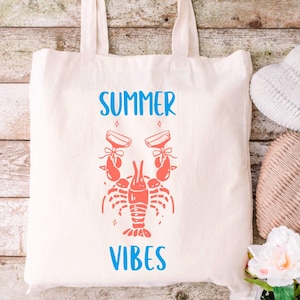 May include: A white tote bag with blue text that says "Summer Vibes". A red lobster with two glasses of champagne is in the center of the bag.