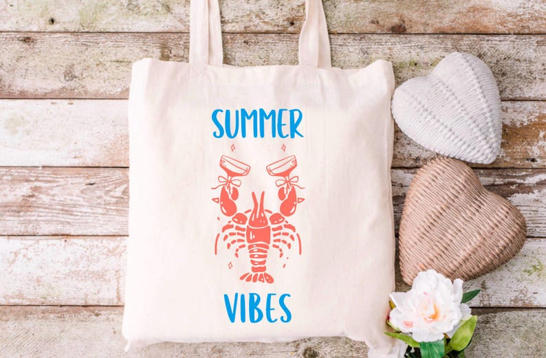 May include: A white tote bag with blue text that says "Summer Vibes". A red lobster with two glasses of champagne is in the center of the bag.