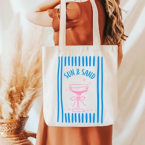 May include: White canvas tote bag with blue stripes and the text "SUN & SAND" in pink. A pink cocktail glass with a blue bow is in the center of the bag.
