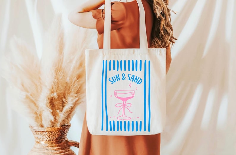 May include: White canvas tote bag with blue stripes and the text "SUN & SAND" in pink. A pink cocktail glass with a blue bow is in the center of the bag.