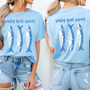 Puede incluir: Camiseta azul claro con la frase "salty but sweet" y un dise&ntilde;o de tres peces azules y blancos. El mismo dise&ntilde;o est&aacute; en la parte delantera y trasera. La modelo lleva unos shorts vaqueros azules claros.