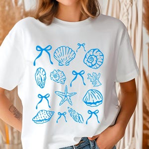 May include: White t-shirt with a blue seashell and starfish design. The design includes various seashells, starfish, and coral.