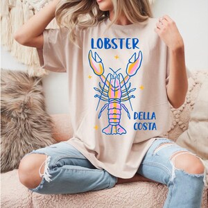 Puede incluir: Camiseta beige con un gr&aacute;fico de langosta colorida. Las palabras "LOBSTER" y "DELLA COSTA" est&aacute;n en azul. La langosta est&aacute; delineada en azul con detalles en rosa, amarillo y naranja. La camiseta se combina con vaqueros azules rotos.