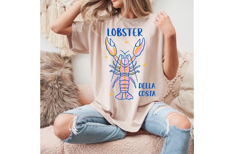 Puede incluir: Camiseta beige con un gr&aacute;fico de langosta colorida. Las palabras "LOBSTER" y "DELLA COSTA" est&aacute;n en azul. La langosta est&aacute; delineada en azul con detalles en rosa, amarillo y naranja. La camiseta se combina con vaqueros azules rotos.