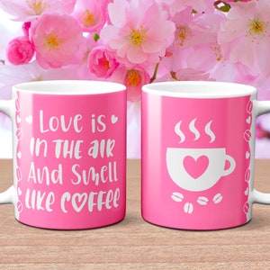 Peut inclure: Deux tasses à café roses avec des poignées blanches. Une tasse a le texte "Love is in the air and smells like coffee" avec un symbole de cœur. L'autre tasse a une tasse à café blanche avec un cœur à l'intérieur et de la vapeur qui sort du haut. Les deux tasses ont une bordure blanche avec des cœurs et des grains de café.