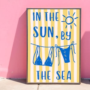 Puede incluir: Un p&oacute;ster enmarcado con un fondo a rayas amarillas y blancas. El p&oacute;ster presenta texto azul que dice "IN THE SUN, BY THE SEA" con un dibujo de un sol y dos bikinis azules colgando de un tendedero.