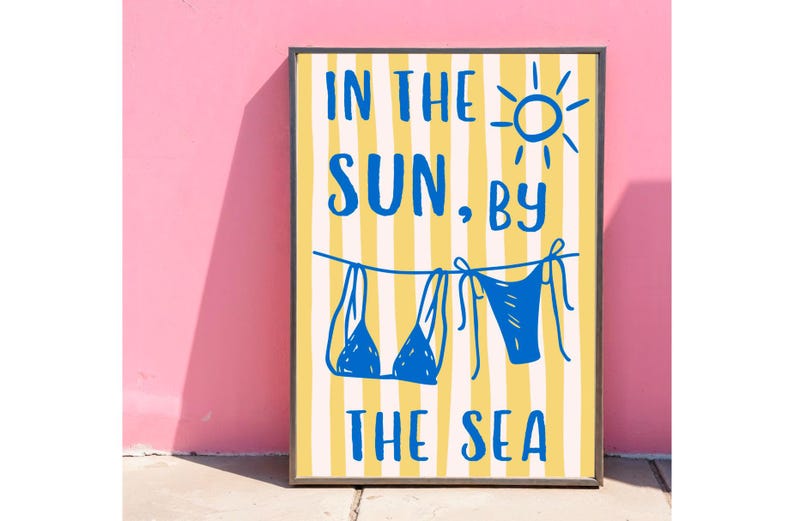 Puede incluir: Un p&oacute;ster enmarcado con un fondo a rayas amarillas y blancas. El p&oacute;ster presenta texto azul que dice "IN THE SUN, BY THE SEA" con un dibujo de un sol y dos bikinis azules colgando de un tendedero.