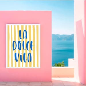 Puede incluir: Una impresi&oacute;n art&iacute;stica enmarcada con las palabras "LA DOLCE VITA" en escritura azul sobre un fondo a rayas amarillas y blancas. La impresi&oacute;n est&aacute; montada en una pared rosa, con una vista del oc&eacute;ano y el cielo al fondo.