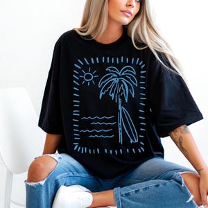 Puede incluir: Camiseta negra con un dise&ntilde;o azul de una palmera, una tabla de surf, un sol y olas. El dise&ntilde;o est&aacute; enmarcado por un rect&aacute;ngulo punteado. La camiseta se lleva con vaqueros azules.