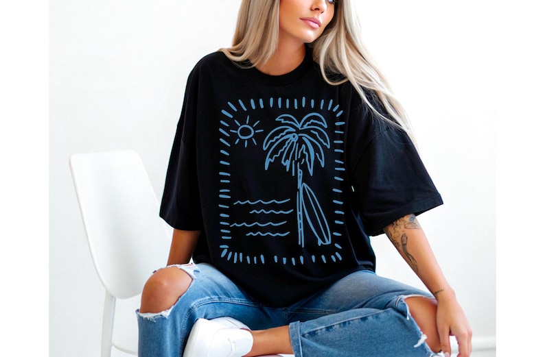 Puede incluir: Camiseta negra con un dise&ntilde;o azul de una palmera, una tabla de surf, un sol y olas. El dise&ntilde;o est&aacute; enmarcado por un rect&aacute;ngulo punteado. La camiseta se lleva con vaqueros azules.
