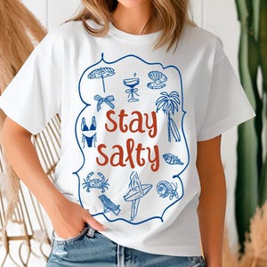 May include: White t-shirt with a blue and red graphic design. The graphic features a variety of summer-themed icons, including a beach umbrella, a cocktail glass, a seashell, a palm tree, a surfboard, a bikini, a crab, and a fish. The text "Stay Salty" is written in red.
