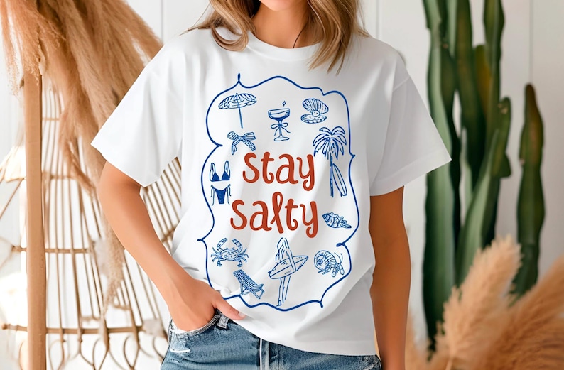 May include: White t-shirt with a blue and red graphic design. The graphic features a variety of summer-themed icons, including a beach umbrella, a cocktail glass, a seashell, a palm tree, a surfboard, a bikini, a crab, and a fish. The text "Stay Salty" is written in red.