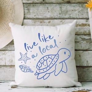 May include: White pillow with blue text that says "Live like a local."  The pillow also features a blue illustration of a sea turtle, a starfish, and a seashell.