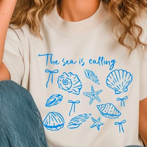 May include: A white t-shirt with a blue graphic that says "The sea is calling" and features various seashells and starfish.