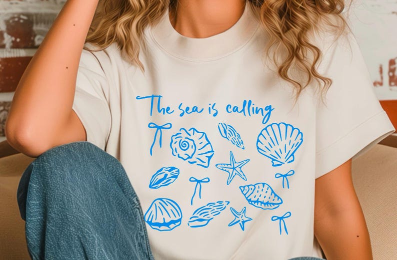 May include: A white t-shirt with a blue graphic that says "The sea is calling" and features various seashells and starfish.