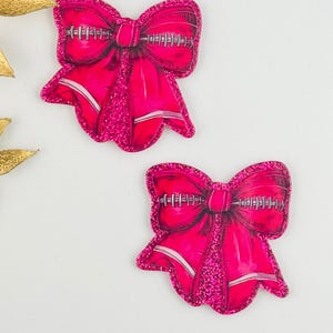 May include: Two bright pink bow-shaped patches featuring a central football lace graphic. Each bow has a glittery texture along the edges and center, with a smooth, shaded pink finish on the loops and tails, creating a dimensional appearance.