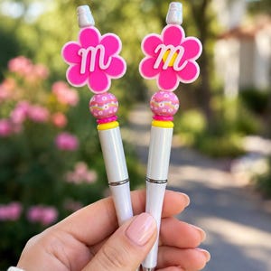 May include: Two white pens featuring a decorative topper shaped like a bright pink flower with a white letter m in the center. Below the flower is a round bead with a colorful wavy pattern and a small yellow spacer ring on the white pen barrel.