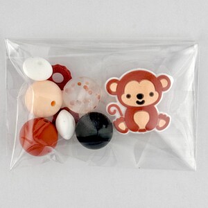 May include: A clear plastic bag containing various beads and a monkey-shaped embellishment. The beads are in shades of white, peach, red, and black. The monkey is brown with a light brown face and ears.
