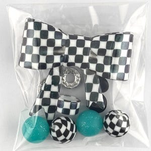 May include: A collection of craft beads and embellishments in a clear plastic bag. The assortment includes a large black and white checkered bow, turquoise and checkered round beads, and silver-colored metal accents.