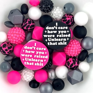 May include: A collection of beads in various shapes, sizes, and colors. The beads include pink, black, white, and gray, with some featuring a leopard print design. Two circular beads display the text: "I don't care + how you + were raised + Unlearn + that shit."