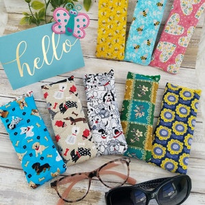 May include: Assortment of colorful fabric eyeglass cases with various patterns, including dogs, bees, flowers, and plaid. The cases are lying on a light wood surface with a pair of reading glasses and sunglasses.