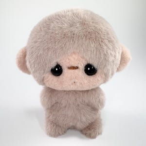 May include: A plush toy featuring a round, fuzzy, light brown head with large, dark, glossy eyes and small ears. The body is covered in soft, textured fur, with a small, horizontal brown stitch detail above the eyes and a tiny dot for a mouth.
