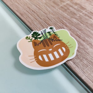 Pode incluir: Um gato castanho com um sorriso branco e plantas verdes a crescerem da sua cabe&ccedil;a. O gato est&aacute; sobre um fundo rosa e verde claro.