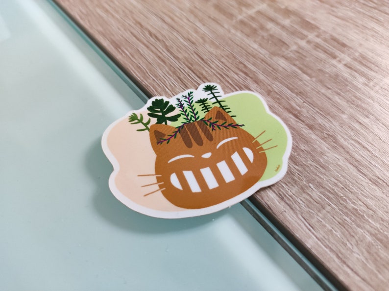 Pode incluir: Um gato castanho com um sorriso branco e plantas verdes a crescerem da sua cabe&ccedil;a. O gato est&aacute; sobre um fundo rosa e verde claro.