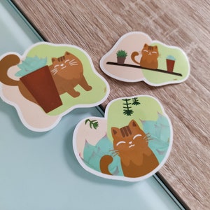 Pode incluir: Tr&ecirc;s adesivos com um gato laranja de desenho animado com listras pretas. O gato &eacute; mostrado em diferentes poses, incluindo sentado em uma prateleira com uma planta em vaso, sentado ao lado de uma suculenta em vaso e relaxando em uma cama de folhas verdes.