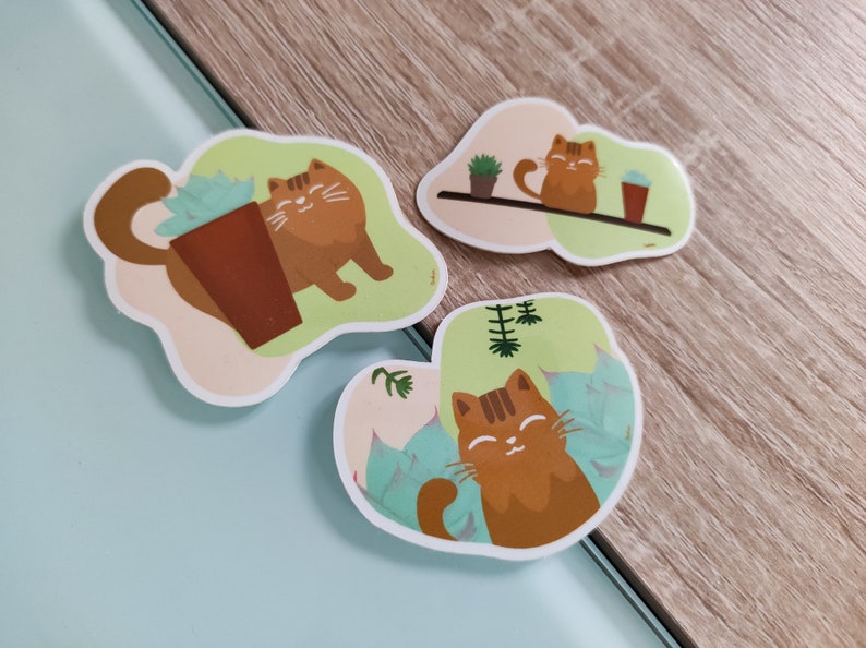 Pode incluir: Tr&ecirc;s adesivos com um gato laranja de desenho animado com listras pretas. O gato &eacute; mostrado em diferentes poses, incluindo sentado em uma prateleira com uma planta em vaso, sentado ao lado de uma suculenta em vaso e relaxando em uma cama de folhas verdes.