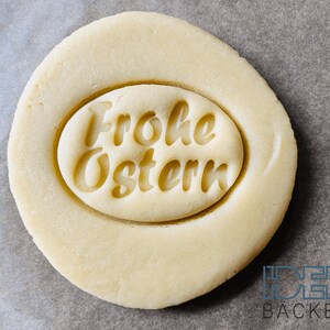 May include: A close-up of a round, light brown cookie dough with a circular imprint that reads "Frohe Ostern" in a cursive font.