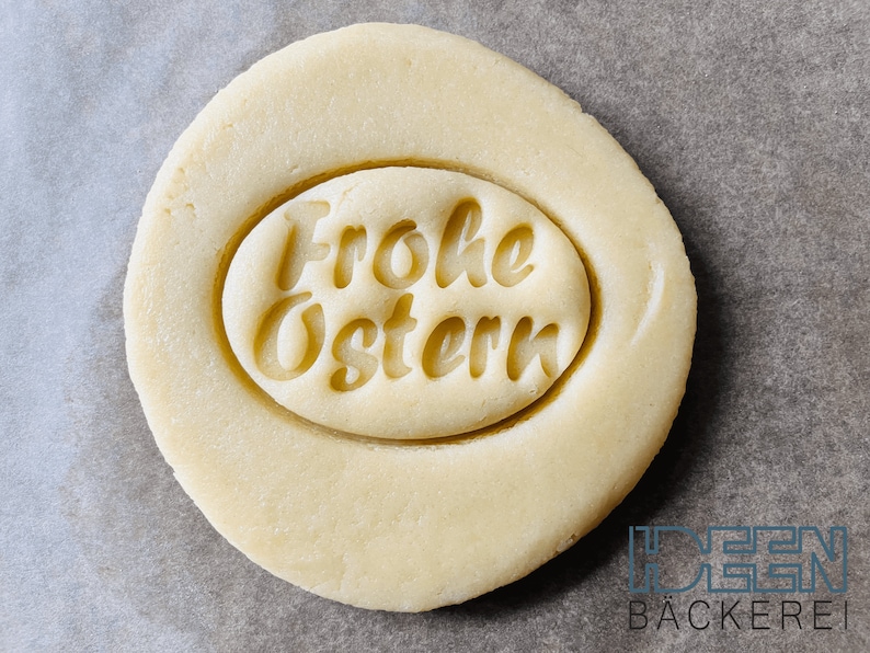 May include: A close-up of a round, light brown cookie dough with a circular imprint that reads "Frohe Ostern" in a cursive font.