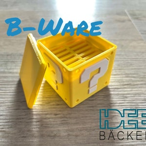 Könnte beinhalten: Eine gelbe 3D-gedruckte Box mit einem Fragezeichen auf der Vorderseite und einem Deckel. Die Box ist mit "B-Ware" und "Ideen Bäckerei" beschriftet.