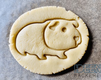 May include: A round, unbaked cookie dough cutout in the shape of a guinea pig. The dough is light beige, with the guinea pig's features clearly defined. The cookie is on a light gray surface.