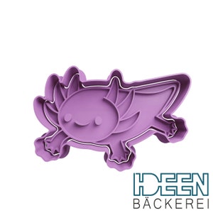 Op de afbeelding: Paarse plastic koekjesvormer in de vorm van een axolotl, een soort salamander. De axolotl heeft een glimlachend gezicht en gevederde kieuwen.