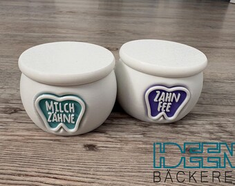 May include: Two white ceramic jars with lids, each featuring a tooth-shaped emblem. One jar has "MILCH ZAHNE" in teal, the other "ZAHN FEE" in purple. The jars are on a wooden surface.