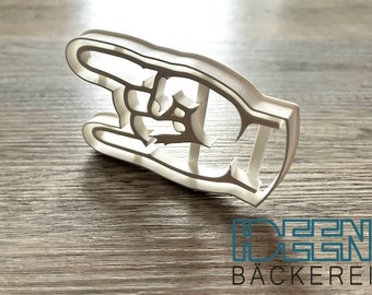 May include: A white plastic cookie cutter in the shape of a hand making a gesture. The cutter is on a wooden surface. The text "BÄCKEREI" is visible at the bottom.