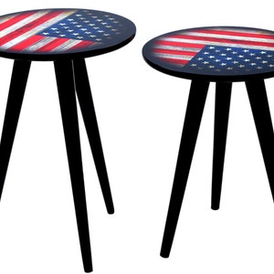 May include: Two small, round side tables. The tabletop features a distressed American flag design with red and white stripes and a blue field of stars. The tables have black, tripod-style legs.
