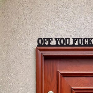 Könnte beinhalten: Schwarzes Metallschild mit der Aufschrift "OFF YOU FUCK" über einer Holztür. Das Schild besteht aus einer einzigen Textzeile, und die Tür hat eine dunkelbraune Farbe.