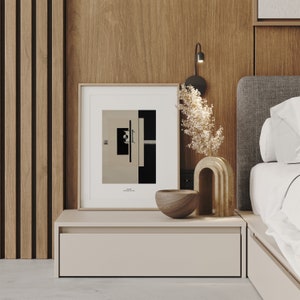 May include: A framed black and white abstract print with a geometric design hangs above a light brown nightstand with a drawer. A wooden bowl and a decorative ceramic sculpture are on the nightstand. The nightstand is next to a bed with a gray headboard and white bedding.