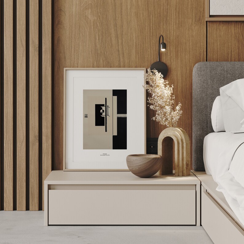 May include: A framed black and white abstract print with a geometric design hangs above a light brown nightstand with a drawer. A wooden bowl and a decorative ceramic sculpture are on the nightstand. The nightstand is next to a bed with a gray headboard and white bedding.