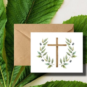May include: A white card with a brown wooden cross surrounded by a green leafy wreath with blue berries. The card is sitting on a brown envelope.