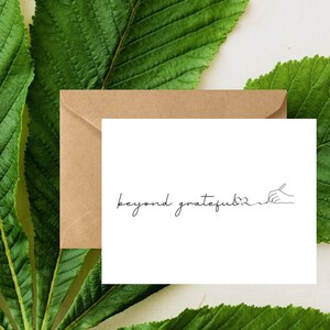 May include: A white card with a brown envelope behind it. The card has the text "beyond grateful" written in black cursive with a heart symbol and a hand drawing a line under the text.