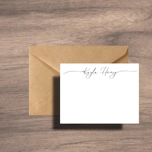 May include: A white stationery card with the name "Kayla Henry" in elegant black script, accompanied by a matching brown envelope. The card and envelope are set against a wooden background, creating a simple and classic aesthetic.
