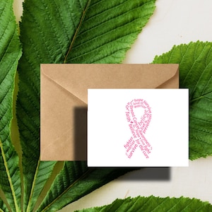 May include: A white card with a pink ribbon design made of words like "hope", "believe", and "survive". A brown envelope sits behind the card, with green leaves in the background. The card is for breast cancer awareness.