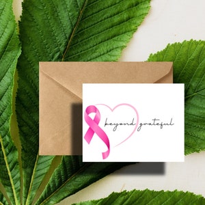 May include: A white card with a pink ribbon and a heart shape with the text "beyond grateful". The card is sitting on a brown envelope and green leaves.